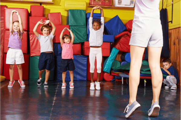 Children stretching their arms up to the ceiling in gym class as they follow along with an instructor.
