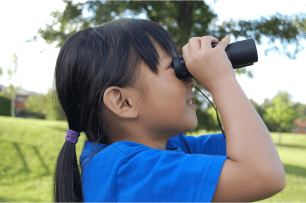 Young girl outside looking up to the sky through a pair of binoculars.