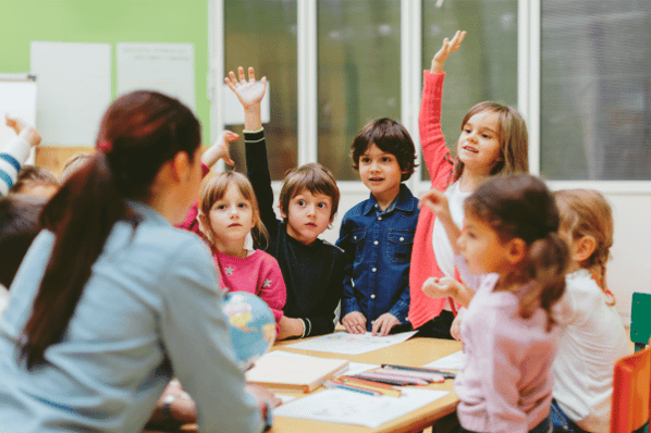 Preschool teacher and children sitting at a table in the classroom. The children are raising their hands.