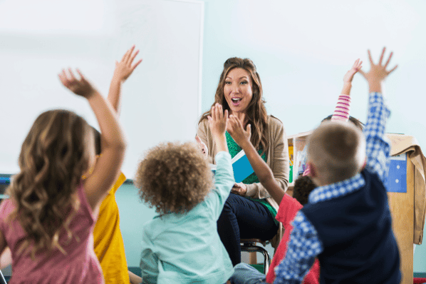 Preschool teacher reading a story to preschool children who are raising their hands.