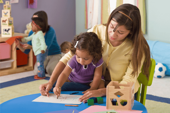 Childcare worker sitting with child at a table helping them to draw a shape with a crayon.