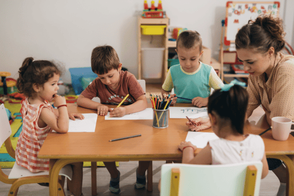 Children sitting at a table at daycare with their teacher. The children are using colored pencils to draw on white pieces of paper as the teacher looks on.