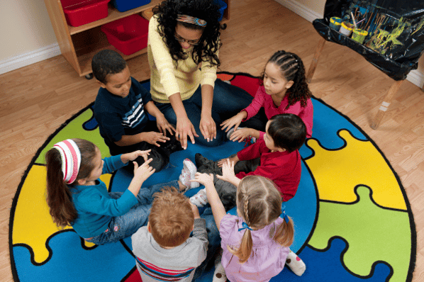 Preschool children sit on a circular rug with their teacher. They are wiggling their fingers in the center of the circle.