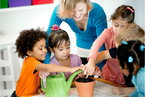 preschool children with teacher gathered around a table, planting seeds in a terracotta pot filled with dirt