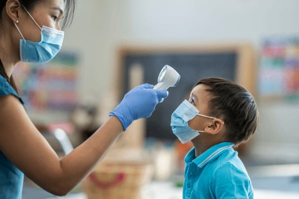 Childcare worker with mask and gloves taking a child's forehead temperature in the classroom.