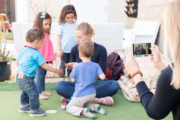 A teacher sits on the ground holding out her hand to show a group of four toddlers around her. In the foreground, another woman uses her phone to take a picture of the scene in front of her.