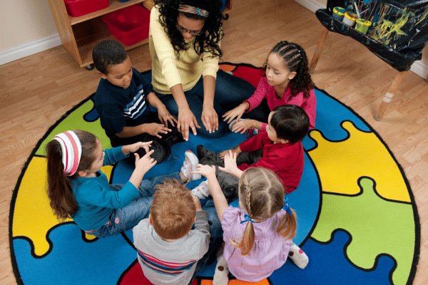 A teacher and six preschool children sitting in a circle on a colorful rug on the floor. They all have their hands and fingers extended into the center of the circle.