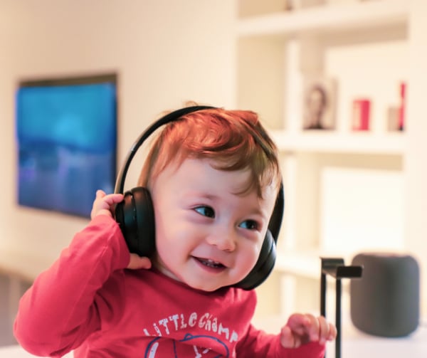 A young child in a red shirt wearing a pair of black headphones and holding one of the earphones against her ear.