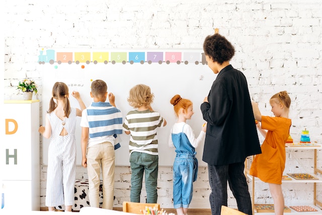 children writing on a whiteboard while teacher observes
