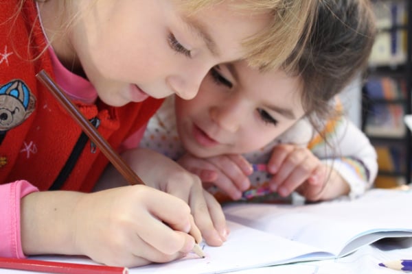 A preschooler in a red shirt leaning over a journal and writing on a blank page with a brown colored pencil. Another preschooler in a white shirt is leaning over and watching as the first preschooler writes.