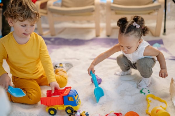 Two toddlers playing with plastic toys in a pile of sand indoors.