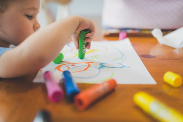 A preschooler holding a green marker and drawing a colorful picture on a sheet of paper. There are orange, blue, and pink markers on the table beside the paper.