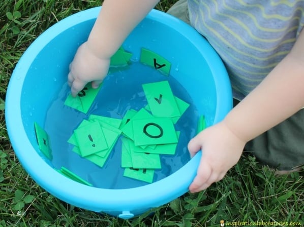 child is kneeling outside on the grass, grabbing green letter cards in a blue bucket filled with water