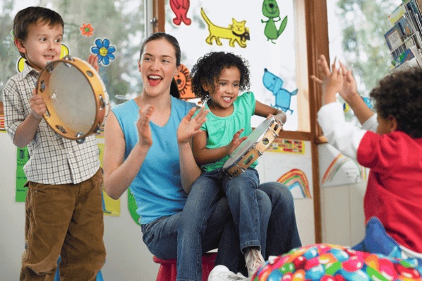 Teacher with children in music class. The children are playing with tambourines as the teacher claps along to the music.