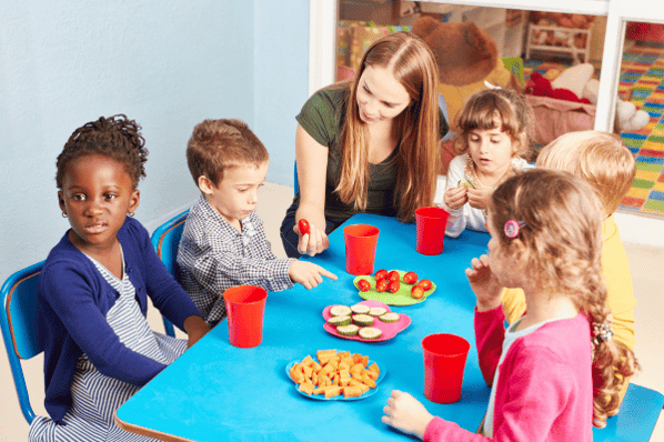 Children in daycare eat vegetables as a snack at the table with their teacher.