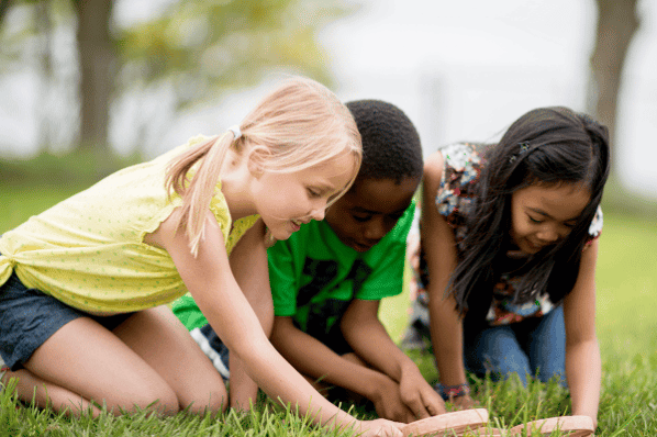 Three children outside using magnifying glasses to look at something in the grass.