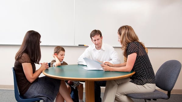 parents, child, and teacher sit at a round table reviewing papers