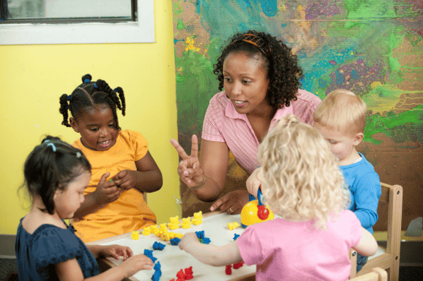 Preschool teacher sitting with four children at a table playing with colorful blocks.
