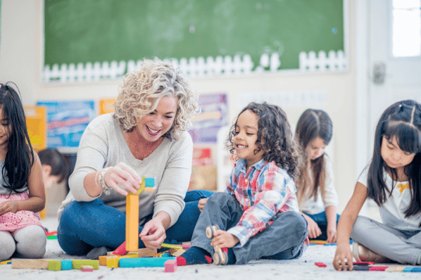 Preschool teacher sitting with child on floor of classroom playing with wooden blocks.