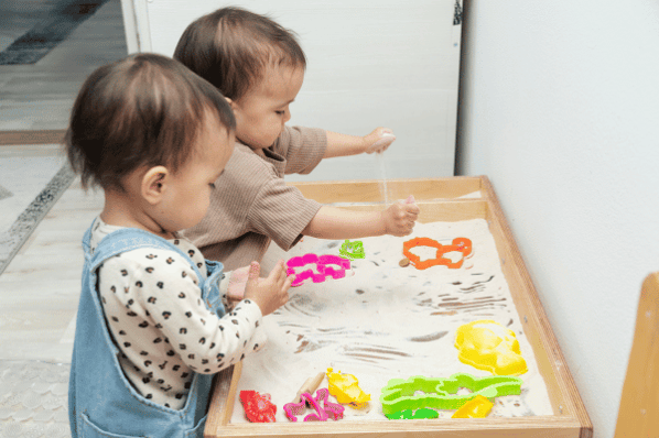 Two toddlers playing with sand and colorful plastic shapes in a sensory bin.
