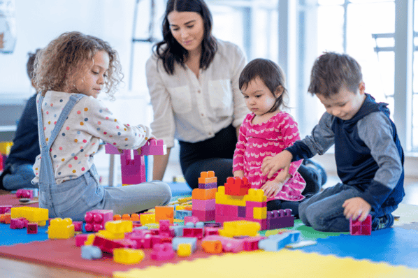 Childcare provider observing as three children build structures with brightly colored legos.