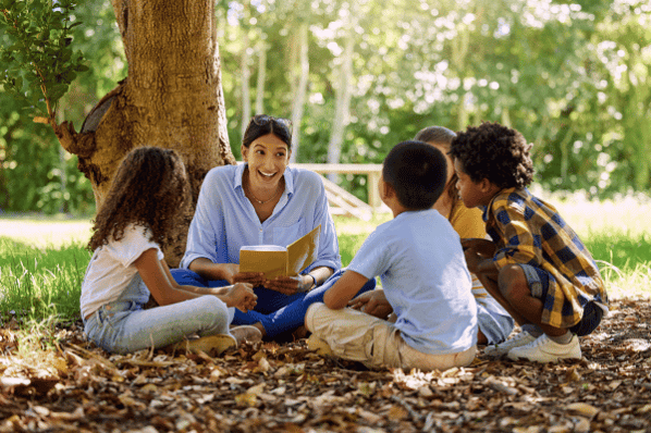 Teacher and students sitting in a circle on the ground outside, underneath a tree. The teacher is reading a book to the children.