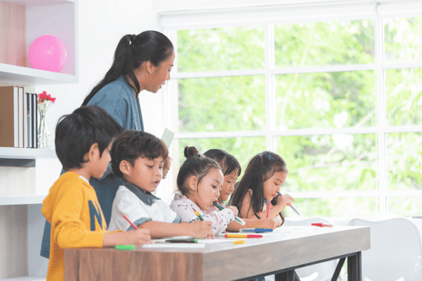 Teacher leans over group of young children at desk, watching them write.