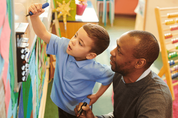 Young boy uses a blue marker to write on a white board, while his male teacher crouches beside him to observe.