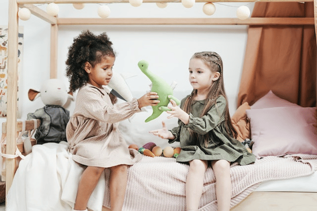 two girls sitting on a bed play holding a dinosaur toy