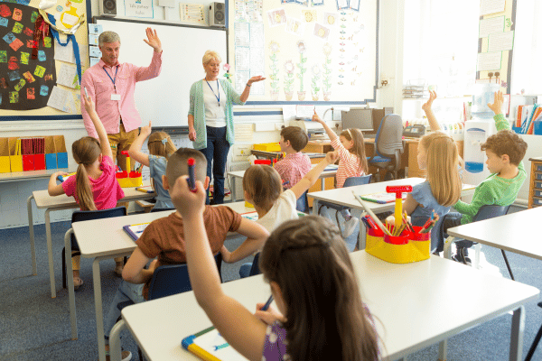 Two teachers stand at the front of a classroom facing children seated at desks.