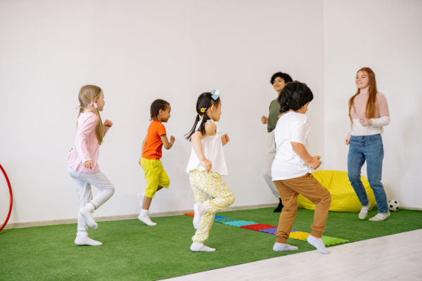 A group of preschoolers and two teachers standing on a green rug and marching in place.