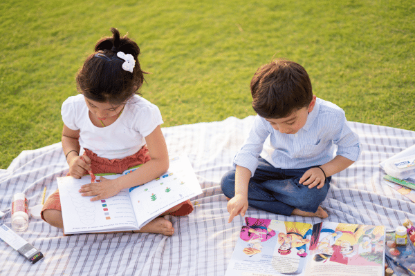Young girl and boy sitting on a checkered blanket on grass outside, both reading picture books.