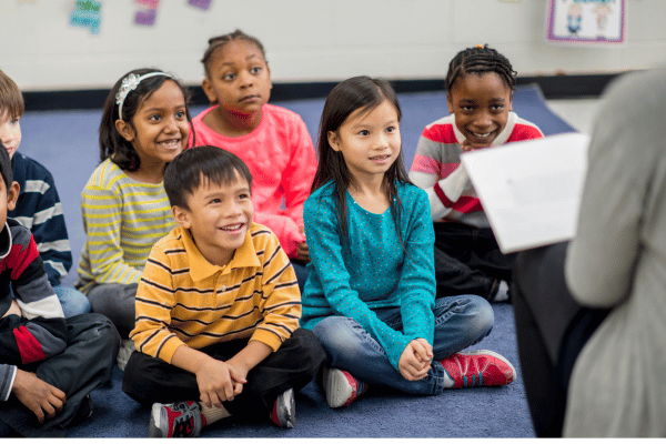 group of preschool children sitting on the ground facing the teacher as she reads from a book