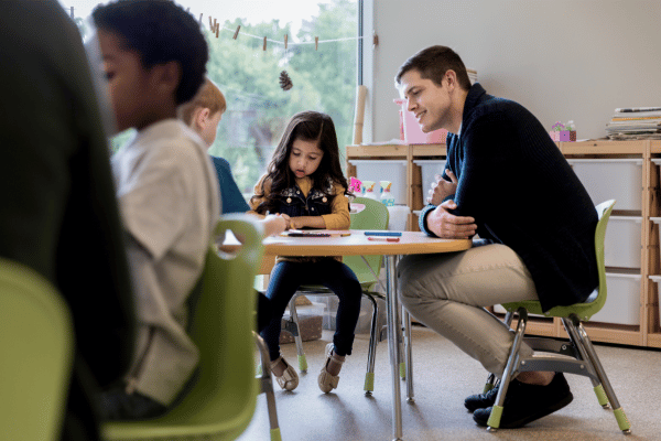 A parent volunteer sits with two children at a table in a preschool classroom