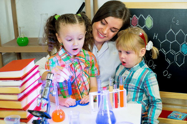 Teacher doing a science experiment with two young children involving water with different colors of food coloring in beakers
