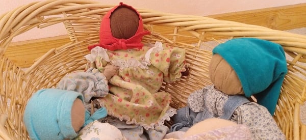 Three cloth dolls of different skin tones in a woven basket. The dolls do not have faces. Two of the dolls are wearing dresses and scarves on their heads. The third doll is wearing a patterned shirt, overalls, and a blue cap.